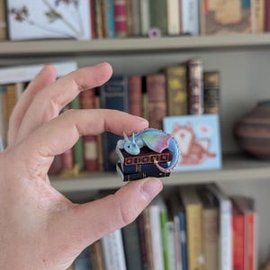 May include: A colorful dragon pin resting on a stack of miniature books. The dragon features blue, pink, and green hues. The books are dark blue with gold lettering. The pin is held in front of a bookshelf filled with various books.