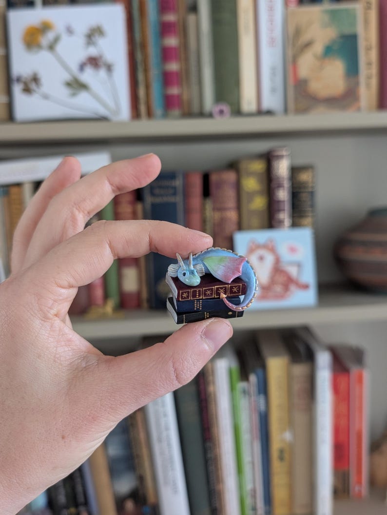 May include: A colorful dragon pin resting on a stack of miniature books. The dragon features blue, pink, and green hues. The books are dark blue with gold lettering. The pin is held in front of a bookshelf filled with various books.