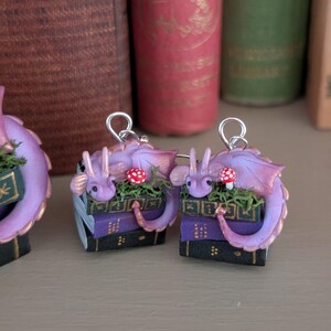 May include: Handmade purple dragon earrings. Each dragon rests on a stack of books with a red and white mushroom. The earrings have a silver-colored hook. The books are purple and black with gold detailing.