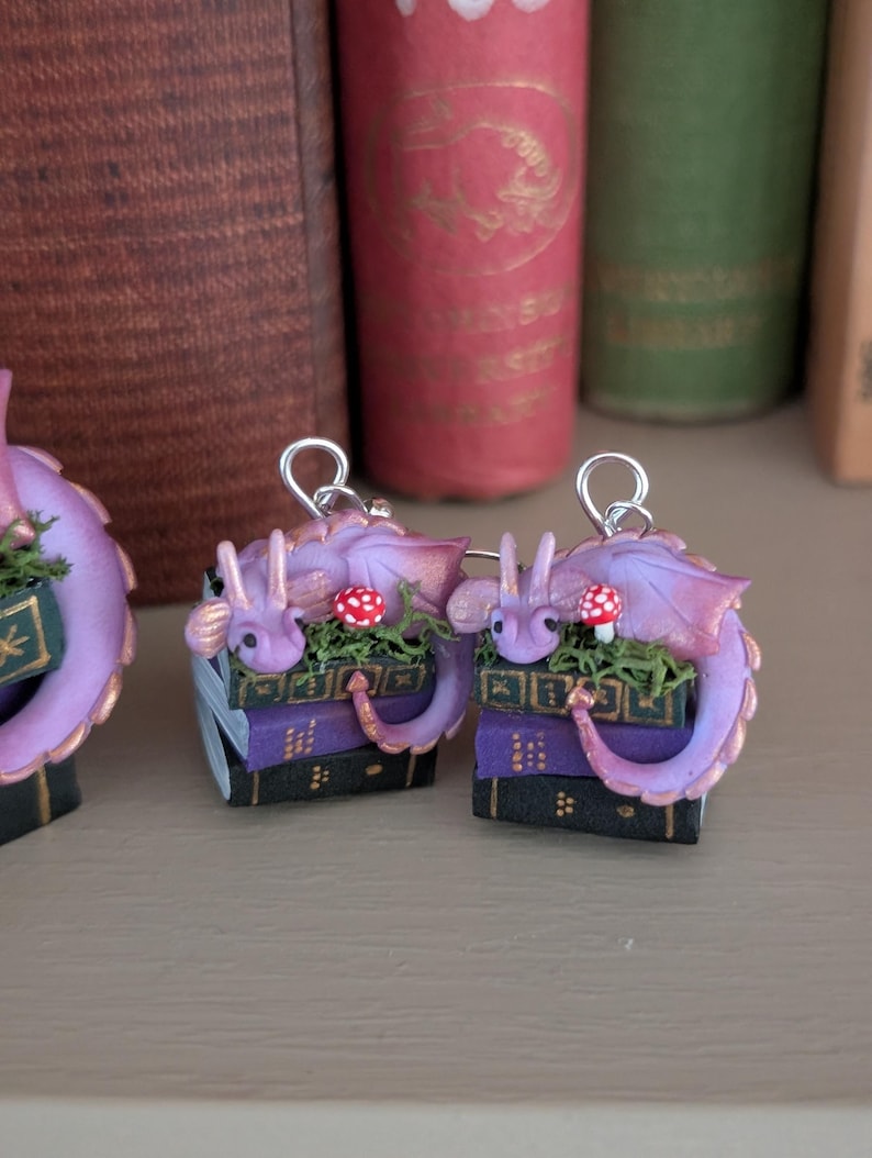 May include: Handmade purple dragon earrings. Each dragon rests on a stack of books with a red and white mushroom. The earrings have a silver-colored hook. The books are purple and black with gold detailing.