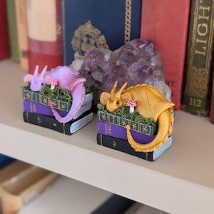Puede incluir: Dos figuras de dragones en miniatura, una morada y otra dorada, descansan sobre pilas de libros. Cada drag&oacute;n est&aacute; posado sobre un lecho de musgo verde con un peque&ntilde;o hongo. Los libros son negros y morados, creando un tema de fantas&iacute;a.