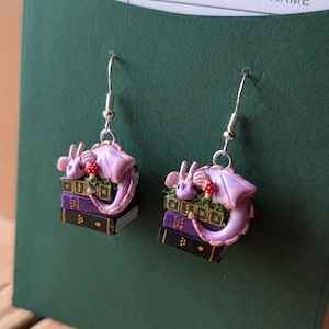 May include: A pair of fantasy-themed earrings. Each earring features a lavender dragon curled around a stack of books. A small red mushroom sits atop the books. The earrings have silver-colored hooks.