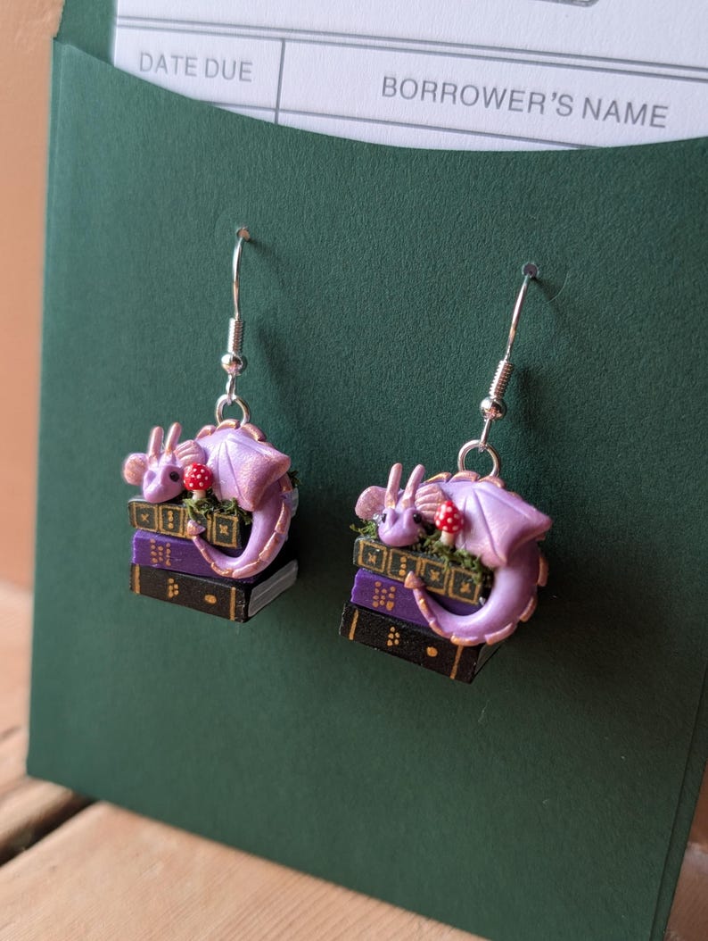 May include: A pair of fantasy-themed earrings. Each earring features a lavender dragon curled around a stack of books. A small red mushroom sits atop the books. The earrings have silver-colored hooks.