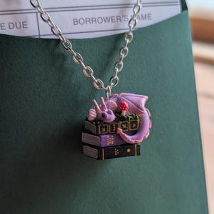 May include: A silver chain necklace with a pendant featuring a purple dragon curled up on a stack of books. The dragon has a red mushroom nearby. The books are black and purple. The necklace is set against a dark green background.