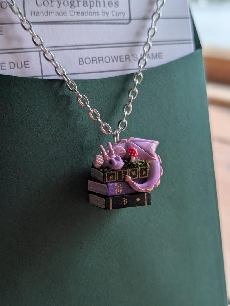 May include: A silver chain necklace with a pendant featuring a purple dragon curled up on a stack of books. The dragon has a red mushroom nearby. The books are black and purple. The necklace is set against a dark green background.