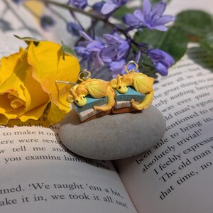 May include: Handmade earrings with a fantasy theme. Each earring features a yellow dragon figurine sitting on a small blue and tan book. The earrings have gold-coloured hooks. The background includes flowers and an open book.