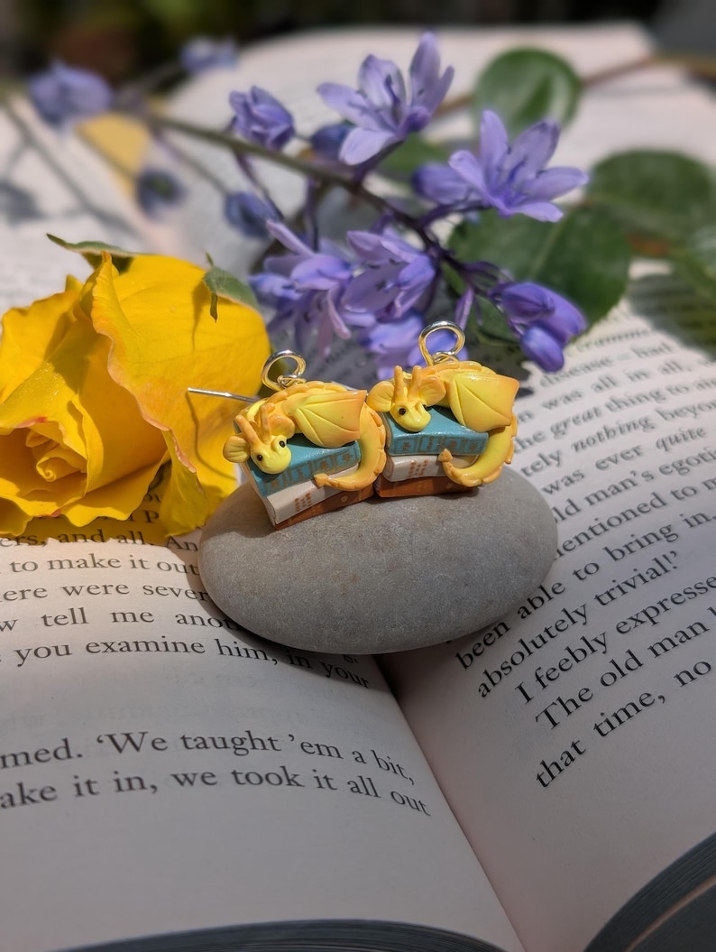 May include: Handmade earrings with a fantasy theme. Each earring features a yellow dragon figurine sitting on a small blue and tan book. The earrings have gold-coloured hooks. The background includes flowers and an open book.