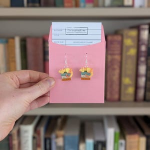 May include: Earrings depicting a small stack of books with a cat, presented in a pink library card pocket. The card reads "Coryographies Handmade Creations by Cory". A bookshelf filled with books forms the backdrop.