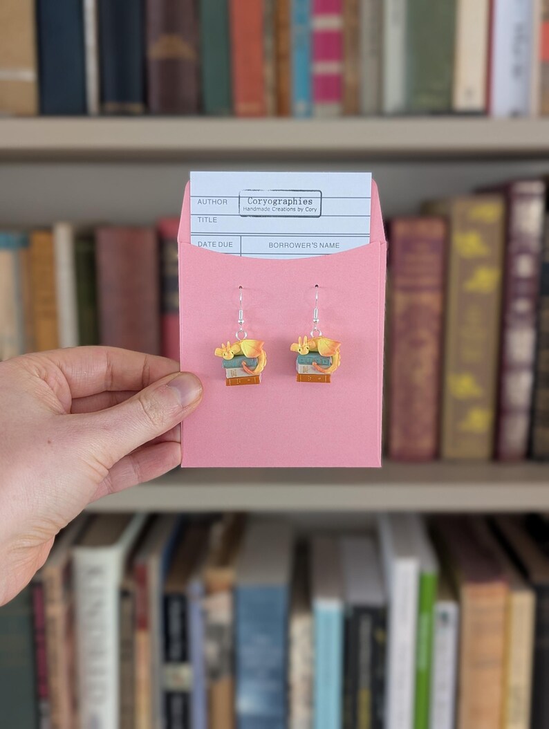 May include: Earrings depicting a small stack of books with a cat, presented in a pink library card pocket. The card reads "Coryographies Handmade Creations by Cory". A bookshelf filled with books forms the backdrop.