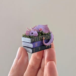 Puede incluir: Una figura de drag&oacute;n p&uacute;rpura en miniatura descansando sobre una pila de tres libros diminutos. El drag&oacute;n tiene alas y cuernos rosas, con una peque&ntilde;a seta roja y blanca cerca. Los libros son negros y morados con detalles dorados, sostenidos en una mano.