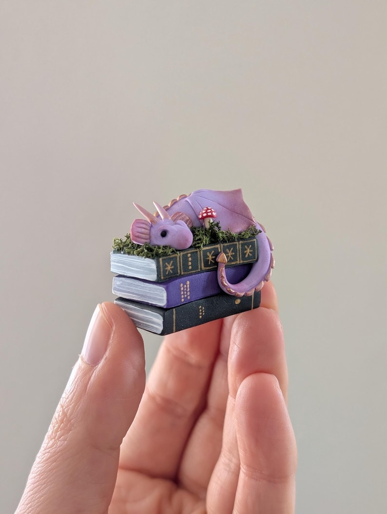 Puede incluir: Una figura de drag&oacute;n p&uacute;rpura en miniatura descansando sobre una pila de tres libros diminutos. El drag&oacute;n tiene alas y cuernos rosas, con una peque&ntilde;a seta roja y blanca cerca. Los libros son negros y morados con detalles dorados, sostenidos en una mano.