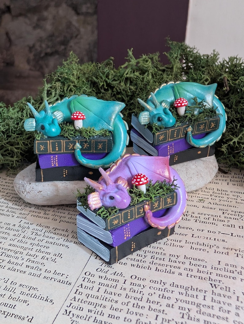 Puede incluir: Tres figuras de dragones en miniatura en tonos azules y morados descansan sobre pilas de libros. Cada drag&oacute;n est&aacute; posado sobre un lecho de musgo verde con una seta roja y blanca. Los libros son negros y morados con detalles dorados.
