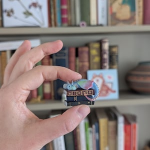 May include: A miniature dragon figurine with blue, green, and pink wings rests atop a stack of tiny books. The books are black with gold detailing. The figurine is held in a person's hand, with a bookshelf in the background.