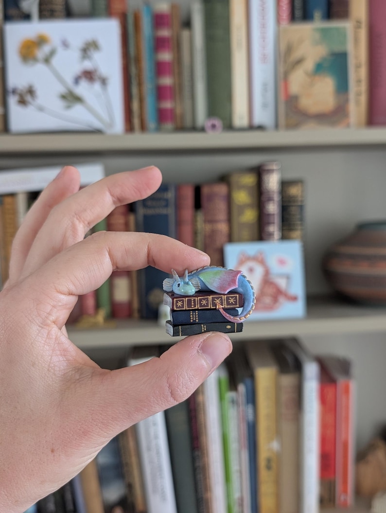 May include: A miniature dragon figurine with blue, green, and pink wings rests atop a stack of tiny books. The books are black with gold detailing. The figurine is held in a person's hand, with a bookshelf in the background.