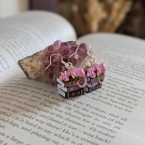 May include: Pink dragon earrings with silver hooks. Each earring features a stack of books with a dragon and mushroom details. The earrings are set against an open book and a purple crystal.