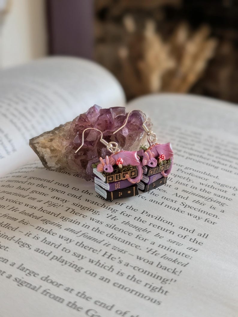 May include: Pink dragon earrings with silver hooks. Each earring features a stack of books with a dragon and mushroom details. The earrings are set against an open book and a purple crystal.