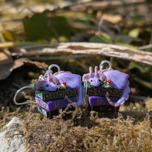May include: A pair of fantasy-themed earrings. Each earring features a small purple dragon resting on a stack of miniature books. The books are purple and black with gold accents. Small red mushroom details are also present.