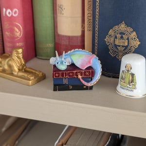 May include: A miniature dragon figurine in blue, pink, and green rests atop a stack of tiny books. The dragon is perched on a shelf with other books and a Shakespeare thimble. The scene evokes a whimsical, fantasy theme.