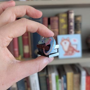 May include: A miniature blue and rainbow-colored dragon figurine perched atop a stack of tiny books. The dragon has a long tail and small wings. The books are dark with gold-colored detailing. The figurine is held in a person's hand.