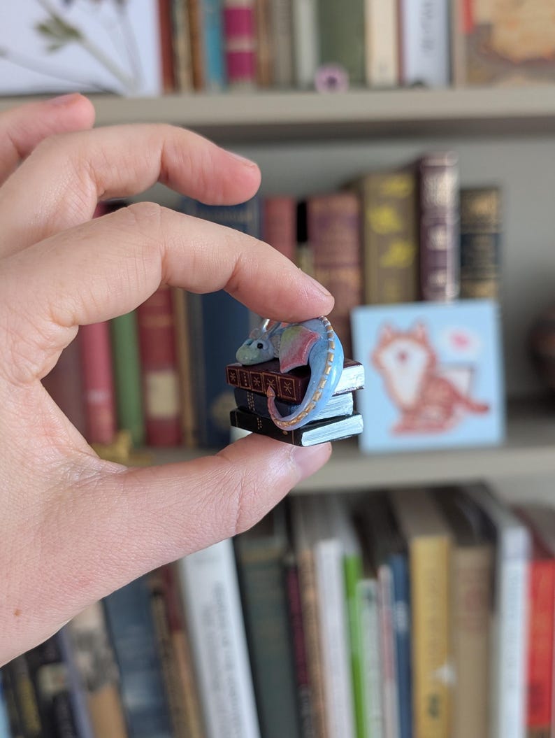 May include: A miniature blue and rainbow-colored dragon figurine perched atop a stack of tiny books. The dragon has a long tail and small wings. The books are dark with gold-colored detailing. The figurine is held in a person's hand.