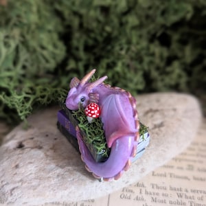 Puede incluir: Figurita artesanal de un drag&oacute;n morado con detalles dorados, posada sobre una pila de libros en miniatura. Una seta roja y blanca est&aacute; cerca. El drag&oacute;n est&aacute; rodeado de musgo verde.