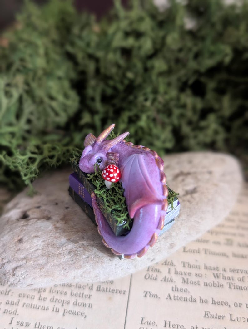 Puede incluir: Figurita artesanal de un drag&oacute;n morado con detalles dorados, posada sobre una pila de libros en miniatura. Una seta roja y blanca est&aacute; cerca. El drag&oacute;n est&aacute; rodeado de musgo verde.