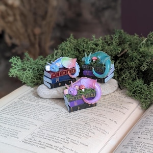 Puede incluir: Tres figuras de dragones en miniatura, cada una posada sobre una pila de libros. Un drag&oacute;n es de color arco&iacute;ris, otro turquesa y el tercero morado. Las figuras est&aacute;n rodeadas de musgo verde y colocadas sobre un libro abierto.