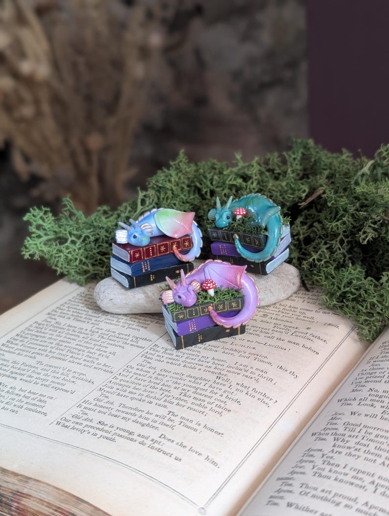 Puede incluir: Tres figuras de dragones en miniatura, cada una posada sobre una pila de libros. Un drag&oacute;n es de color arco&iacute;ris, otro turquesa y el tercero morado. Las figuras est&aacute;n rodeadas de musgo verde y colocadas sobre un libro abierto.
