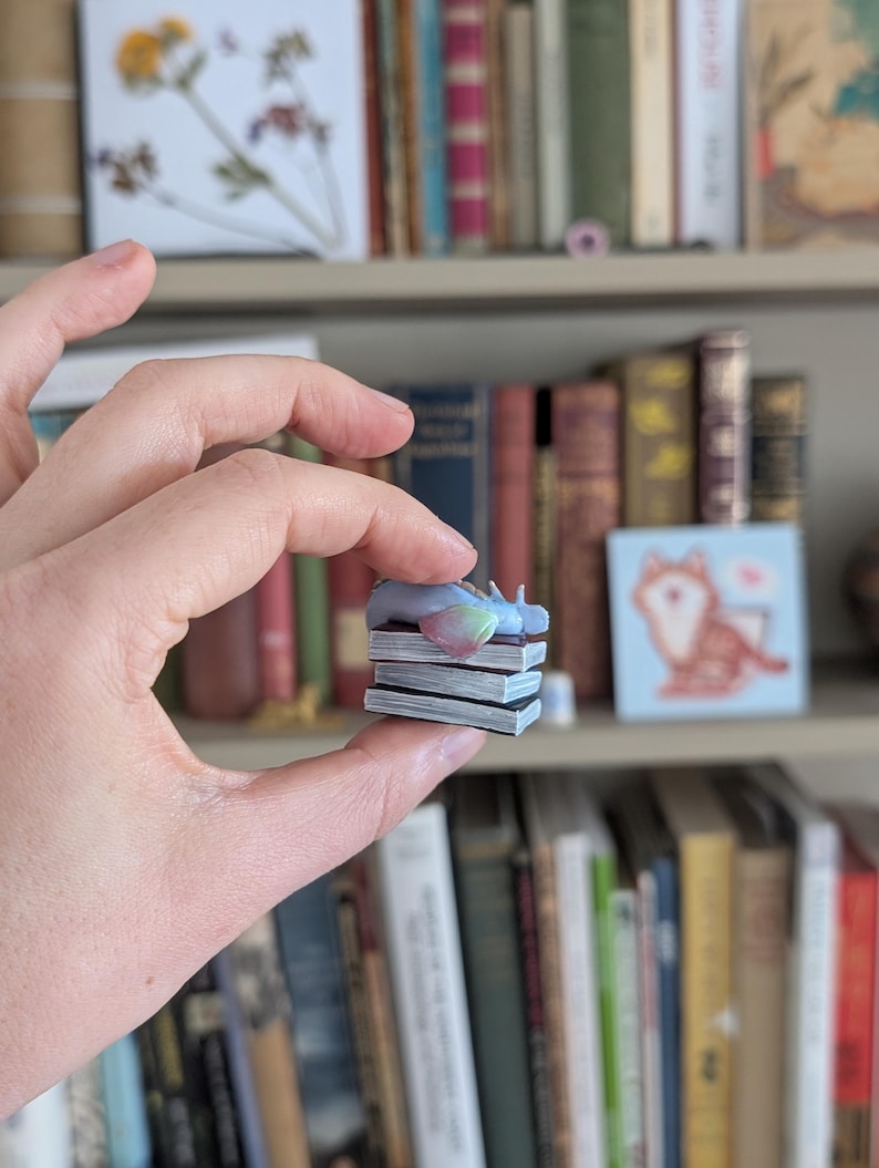 May include: A miniature figurine of a sleeping unicorn with a rainbow-colored mane and tail, resting atop a stack of tiny books. The figurine is held in a hand, with a backdrop of a bookshelf filled with books.