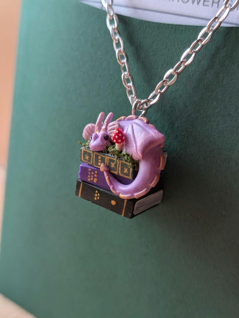 A tiny pendant of a purple dragon with gold detail sits on three books, green, purple and black, which have gold detail on their spines. There is moss around the dragon and a tiny red and white spotted mushroom. It hangs from a silver chain against a dark green background.
