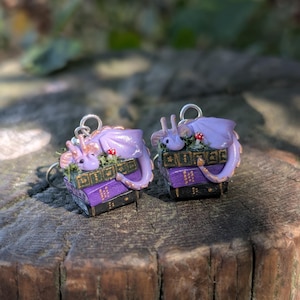 May include: A pair of fantasy-themed earrings. Each earring features a small, lavender dragon curled atop a stack of miniature books. The books are purple and black with gold accents. Small red mushrooms and green foliage add detail. The earrings have silver hooks.