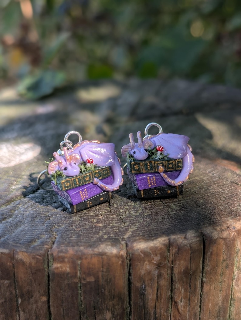 May include: A pair of fantasy-themed earrings. Each earring features a small, lavender dragon curled atop a stack of miniature books. The books are purple and black with gold accents. Small red mushrooms and green foliage add detail. The earrings have silver hooks.