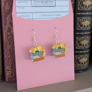May include: Dangle earrings with a yellow and orange dragon design, resting on a stack of books. The earrings hang from silver-coloured hooks and are displayed on a pink card. The card has the text "Coryographies Handmade Creations by Cory".