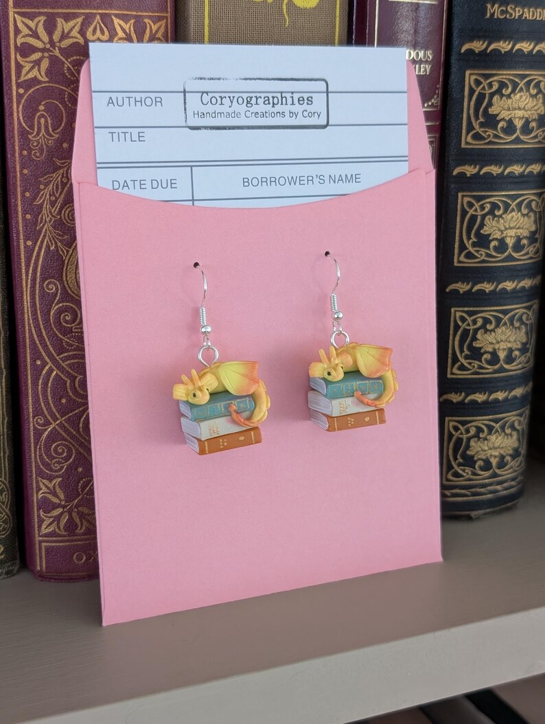 May include: Dangle earrings with a yellow and orange dragon design, resting on a stack of books. The earrings hang from silver-coloured hooks and are displayed on a pink card. The card has the text "Coryographies Handmade Creations by Cory".