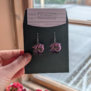 May include: A pair of pink and purple earrings with a unique design, held in a black envelope. The earrings have a detailed, layered appearance with small embellishments. The envelope contains a card with the text "Coryographies Handmade Creations by Cory".