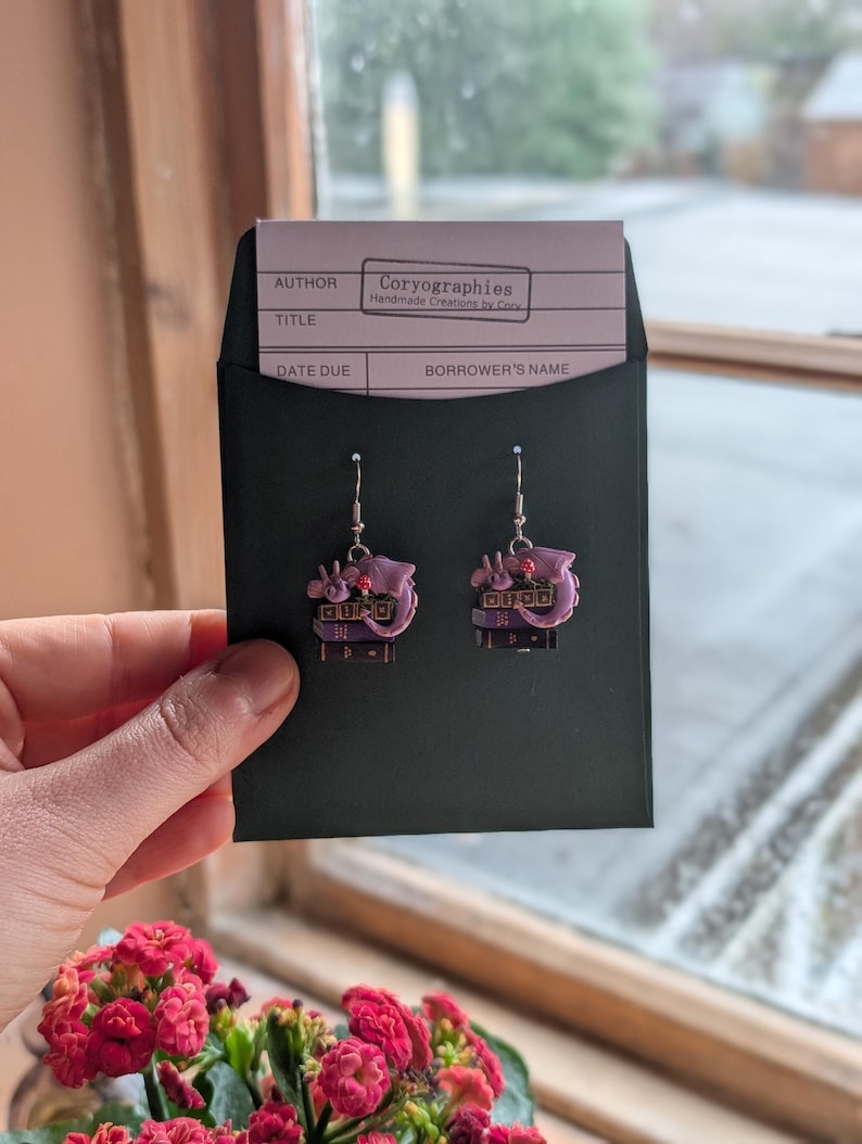 May include: A pair of pink and purple earrings with a unique design, held in a black envelope. The earrings have a detailed, layered appearance with small embellishments. The envelope contains a card with the text "Coryographies Handmade Creations by Cory".