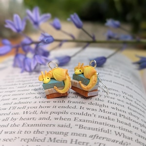 May include: A pair of quirky earrings with yellow dragon figures on miniature book stacks. The dragons have orange details. The earrings are set against a blurred background of a book and purple flowers.