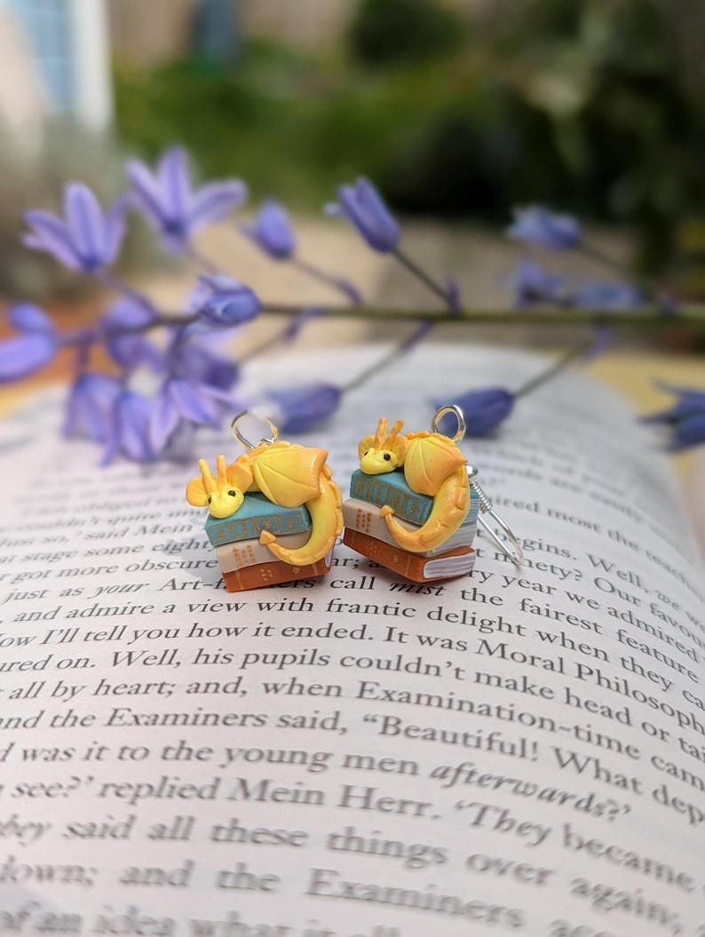 May include: A pair of quirky earrings with yellow dragon figures on miniature book stacks. The dragons have orange details. The earrings are set against a blurred background of a book and purple flowers.