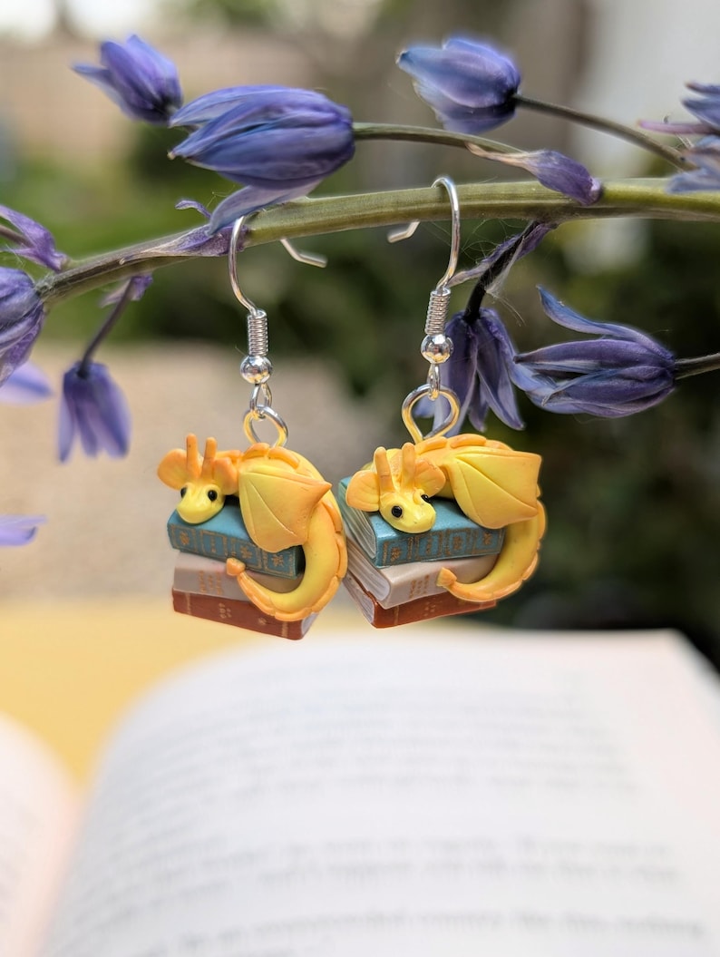 Two earrings dangle from a horizontal stem of bluebells. There are three books, rust, tan and sage, with gold detail on the front. On top of them sit a golden yellow dragon, with their tail wrapped around the front of the books.