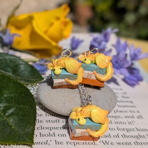 May include: A silver necklace with three pendants. Each pendant features a small, yellow dragon figurine on a stack of miniature books. The background includes flowers and a book page.