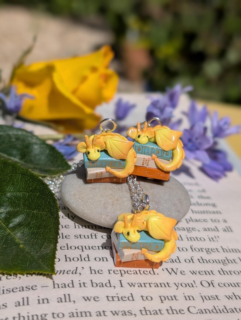 May include: A silver necklace with three pendants. Each pendant features a small, yellow dragon figurine on a stack of miniature books. The background includes flowers and a book page.
