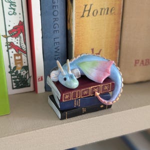 On a book case, a blue dragon lies on three books. it has a green and red wing showing, and gold detail on the horns and scales down it's back. the books are maroon, navy and black, and have gold detail on the spines.