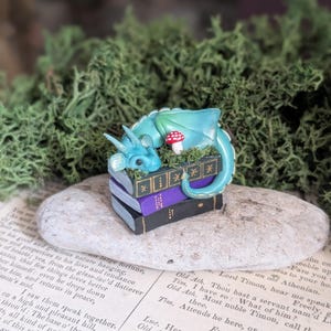 Puede incluir: Una figura caprichosa de un drag&oacute;n turquesa descansando sobre una pila de libros en miniatura. Un hongo rojo y blanco est&aacute; cerca. El drag&oacute;n est&aacute; posado sobre una piedra, rodeado de vegetaci&oacute;n y un fondo de p&aacute;ginas de libros antiguos.