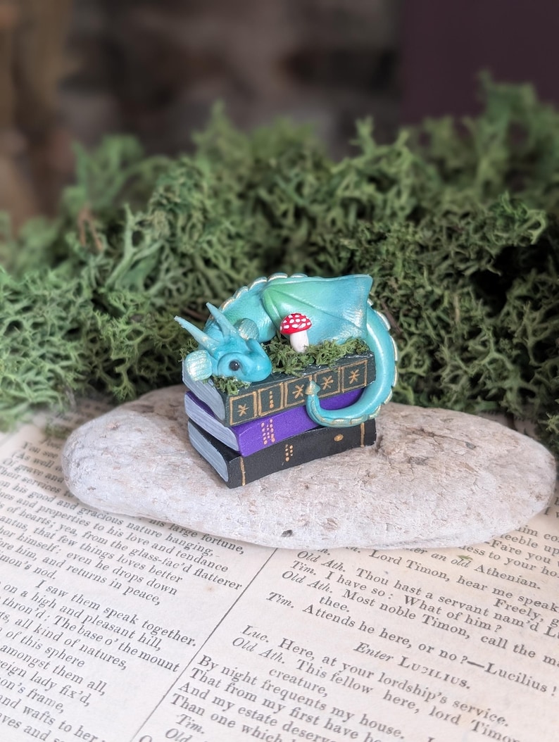Puede incluir: Una figura caprichosa de un drag&oacute;n turquesa descansando sobre una pila de libros en miniatura. Un hongo rojo y blanco est&aacute; cerca. El drag&oacute;n est&aacute; posado sobre una piedra, rodeado de vegetaci&oacute;n y un fondo de p&aacute;ginas de libros antiguos.