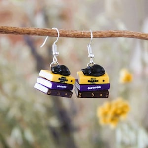 May include: A pair of dangle earrings featuring a stack of three miniature books with a black cat sleeping on top. The books are yellow, purple, and brown.