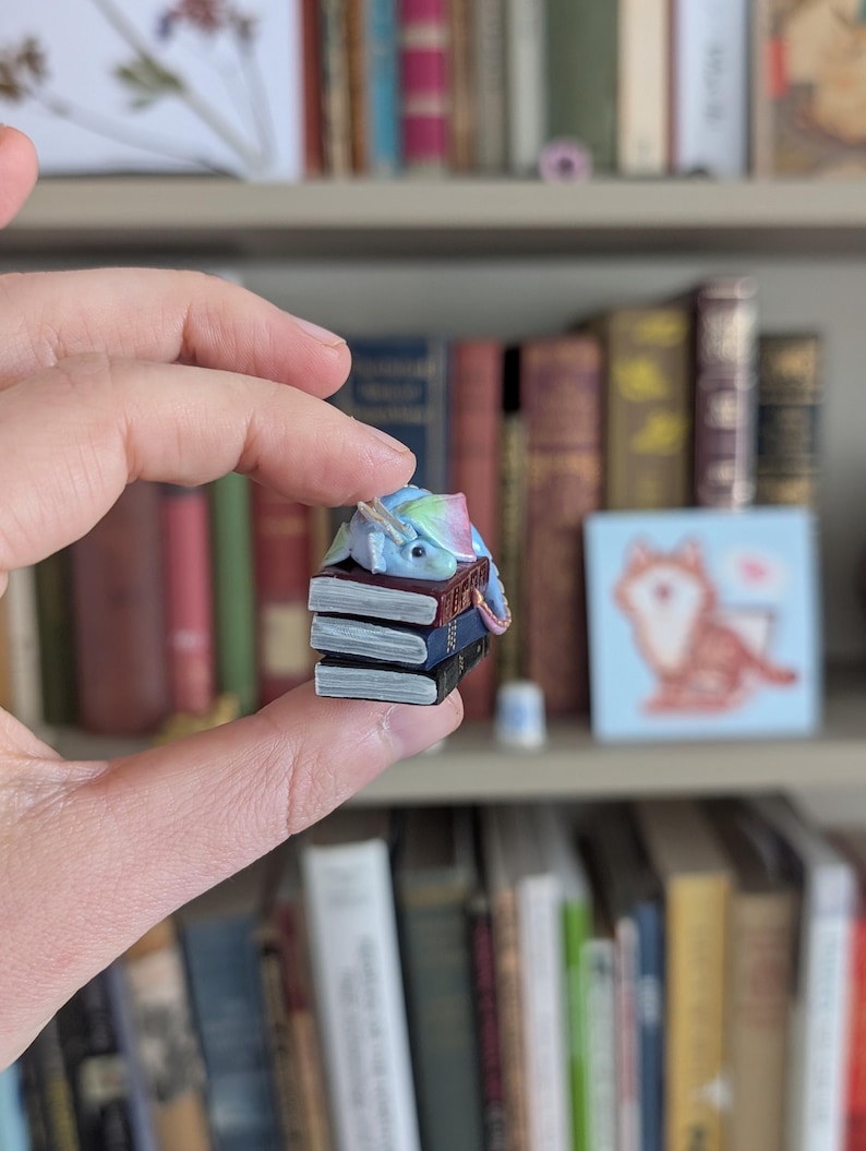 May include: A miniature fantasy figurine of a sleeping unicorn with a blue, pink, and green mane and tail, resting on a stack of tiny books. The books are black, blue, and brown. The figurine is held in front of a bookshelf filled with books.