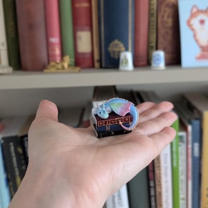 May include: A miniature, colorful dragon figurine resting on a stack of tiny books. The dragon has blue, green, and pink wings and a curled tail. The books are dark with gold lettering. The background includes a bookshelf filled with books.