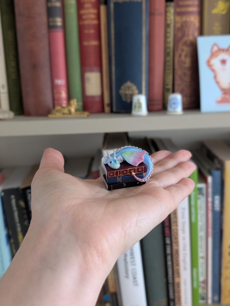 May include: A miniature, colorful dragon figurine resting on a stack of tiny books. The dragon has blue, green, and pink wings and a curled tail. The books are dark with gold lettering. The background includes a bookshelf filled with books.
