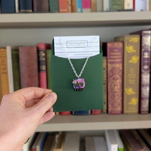 May include: A silver chain necklace with a pendant featuring a purple dragon design. The pendant is displayed on a green card inside a green envelope, held in front of a bookshelf filled with books.