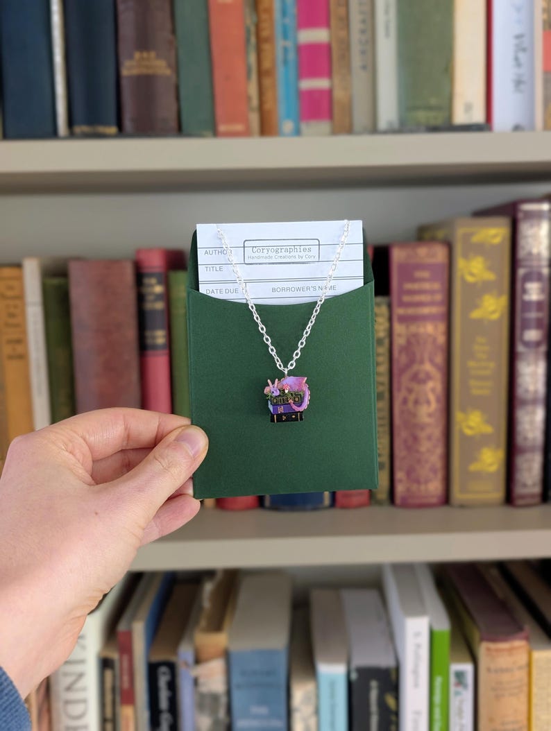 May include: A silver chain necklace with a pendant featuring a purple dragon design. The pendant is displayed on a green card inside a green envelope, held in front of a bookshelf filled with books.
